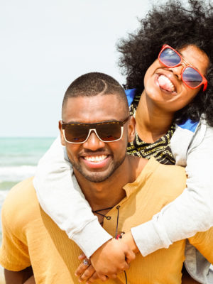 Young afro american couple playing piggyback ride on beach - Cheerful african friends having fun at day with blue ocean background - Concept of lovers happy moments on summer holiday - Vintage filter