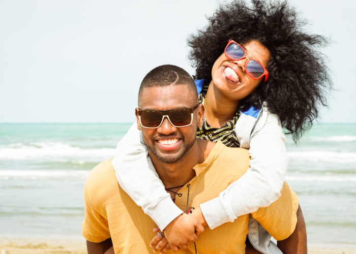 Young afro american couple playing piggyback ride on beach - Cheerful african friends having fun at day with blue ocean background - Concept of lovers happy moments on summer holiday - Vintage filter